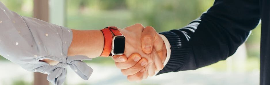 Close-up of a handshake between two adults, one wearing a smartwatch, set in an outdoor environment.